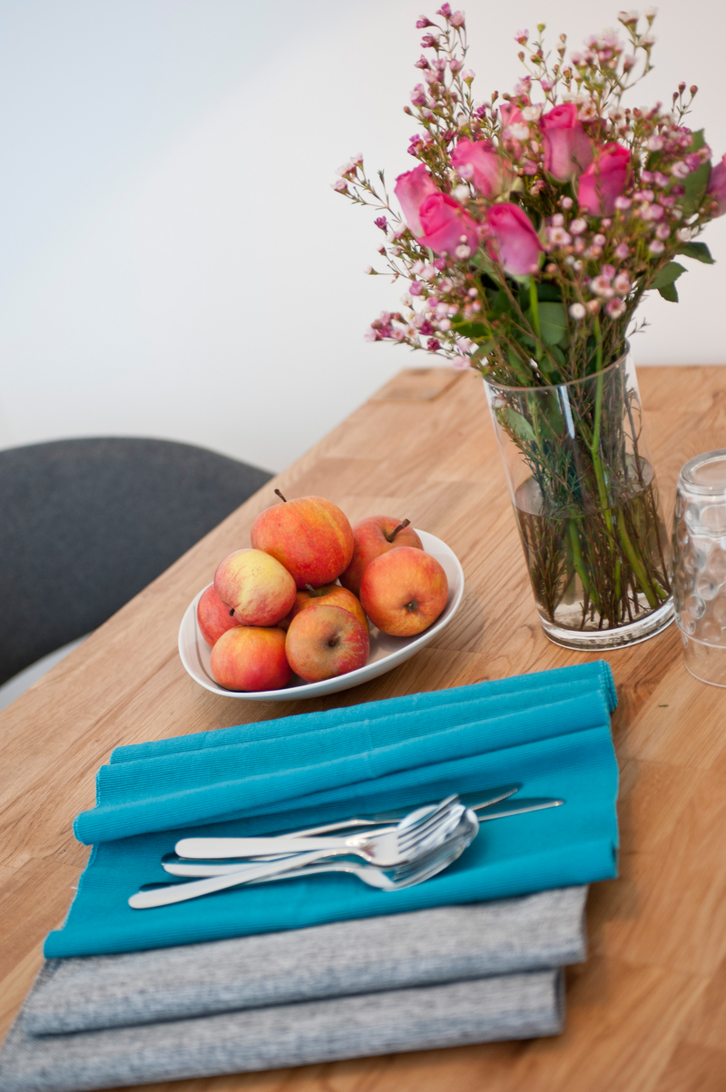 Tisch mit Platzdeckchen und Besteck, Äpfel in einer Schale, Vase mit Blumen.