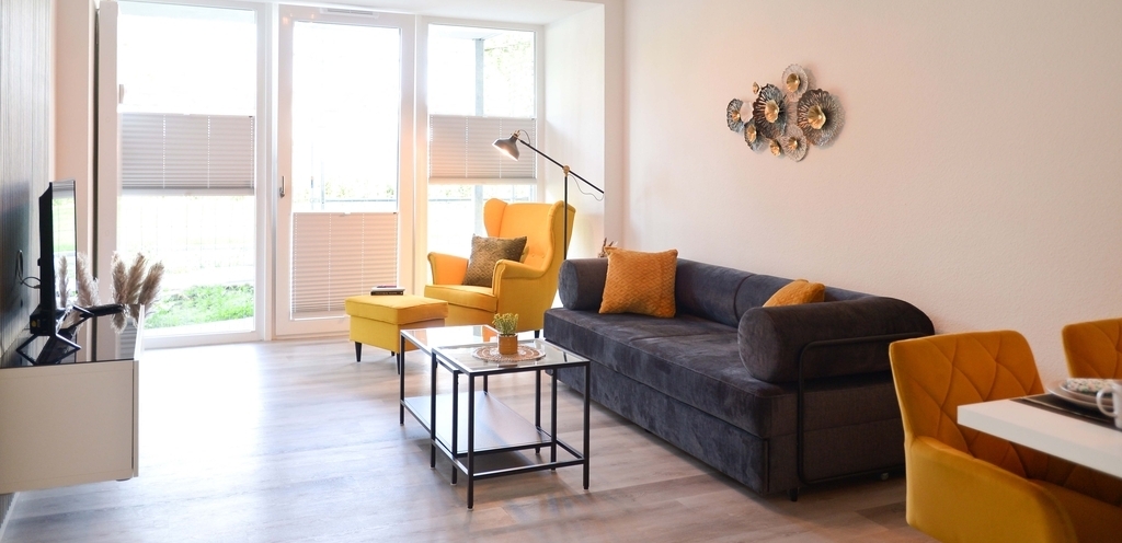 Die Gästewohnung der TRAVE sind vollständig eingerichtet, hier mit gemütlichem Sofa und einem Ohrensessel. Links sieht man einen Flachbildfernseher, rechts eine Essgruppe.