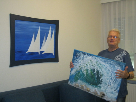 Ein ehrenamtlich Tätiger hält ein selbstgemaltes Bild in der Hand. An der Wand dahinter befindet sich ein weiteres Bild, das im TRAVE-Treff entstanden ist. Ein ehrenamtlich Tätiger hält ein selbstgemaltes Bild in der Hand. An der Wand dahinter befindet sich ein weiteres Bild, das im TRAVE-Treff entstanden ist.