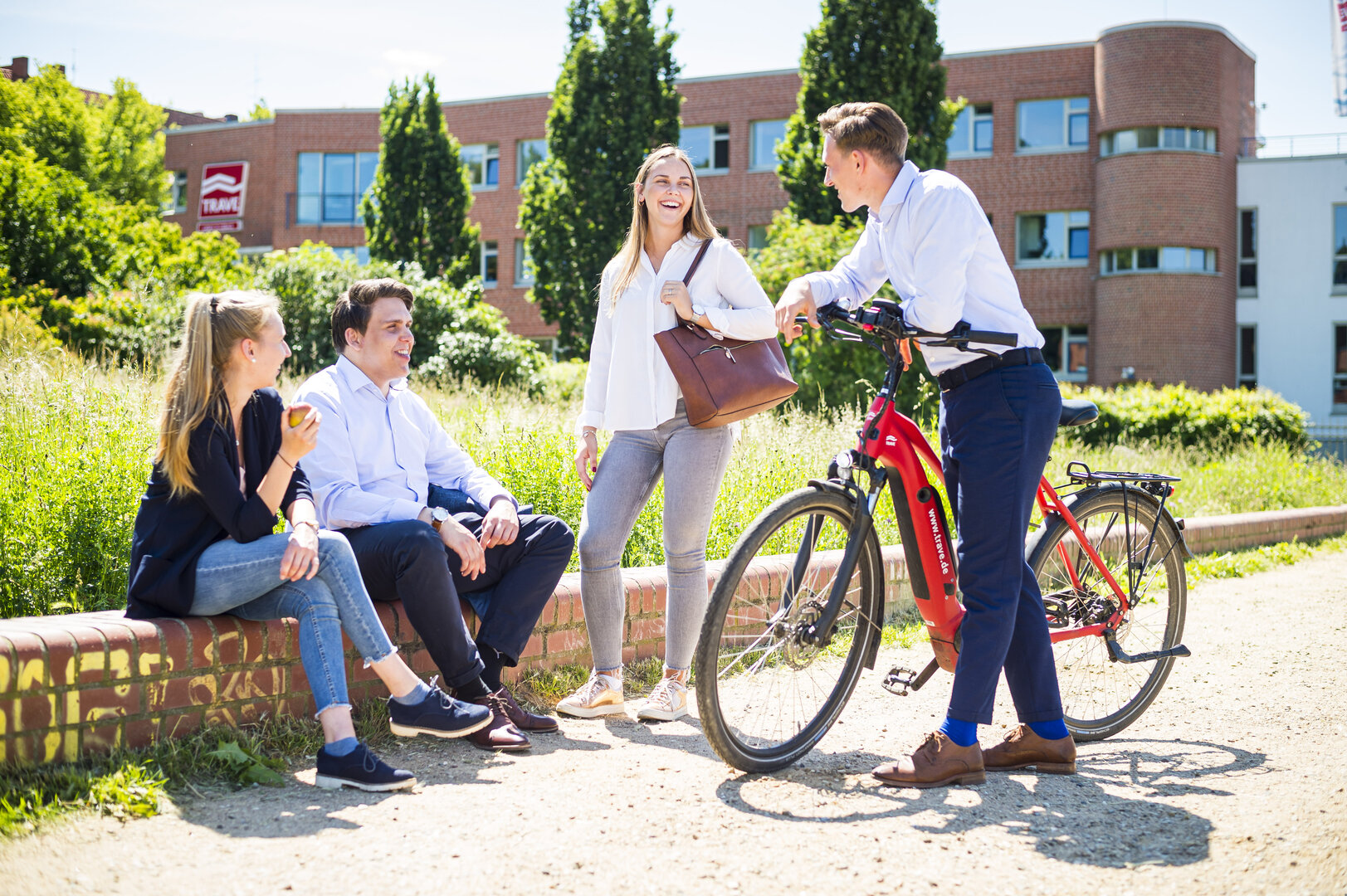 Eine Gruppe von vier Auszubildenden unterhält sich entspannt. Im Hintergrund das Firmengebäude der TRAVE.